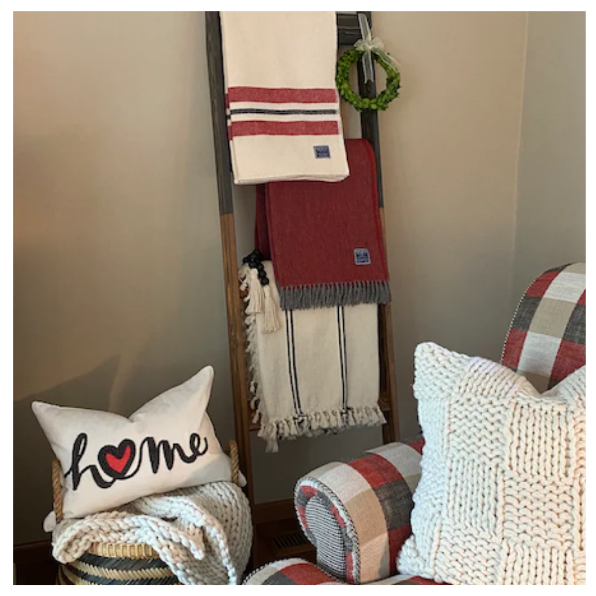 Two tone 6 foot blanket ladder. Top half is stained black and the bottom dark walnut. White and red blankets are hung from the ladder. A plush chair and stool sit next to the ladder, each with pillows a top.