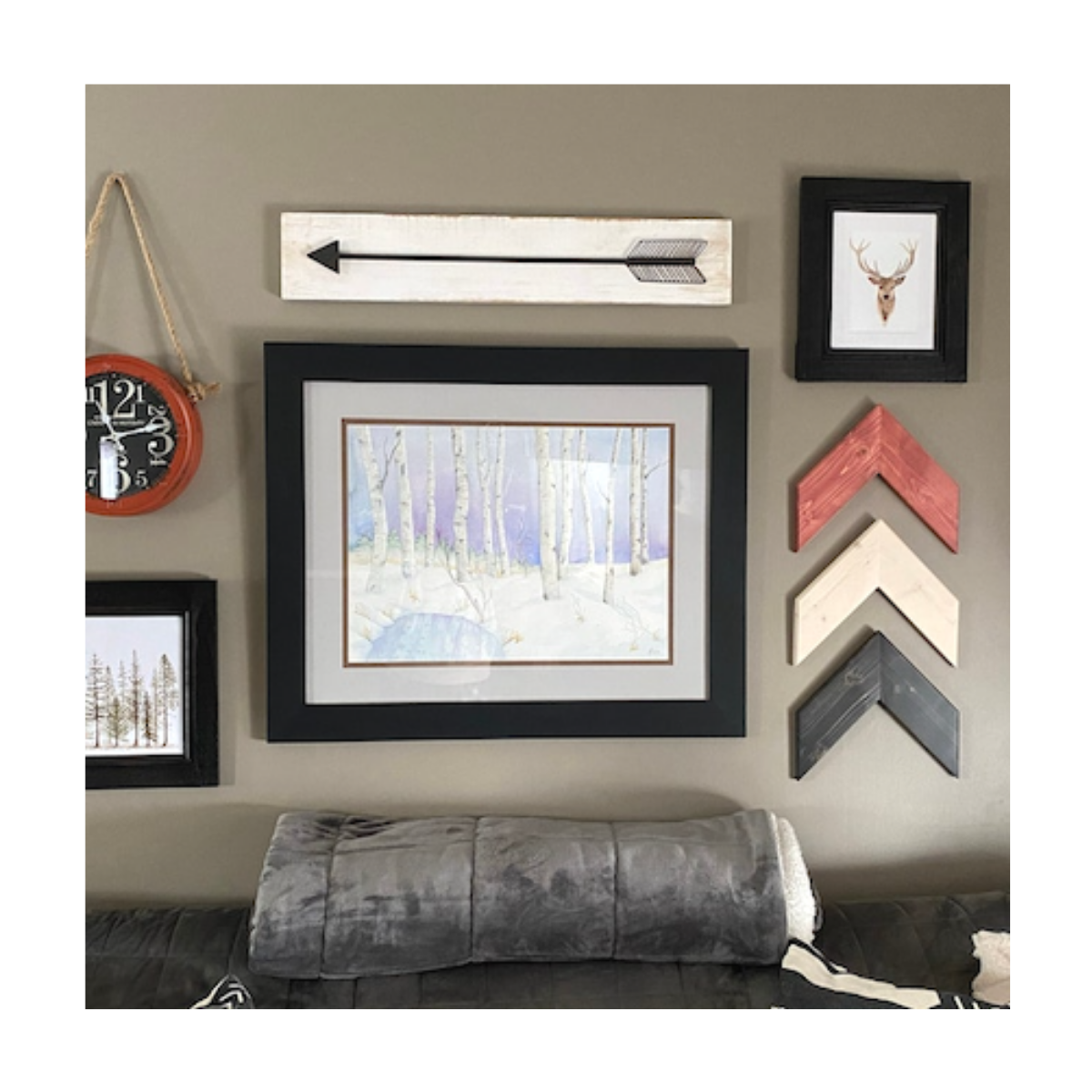 Looking over a couch nested against a wall, several pieces of wall art reside. A black face clock rimed in a red shell. Black wooden arrow on a white background. Three chevron arrows pointing upwards in red, white, and black. Also three framed pictures.